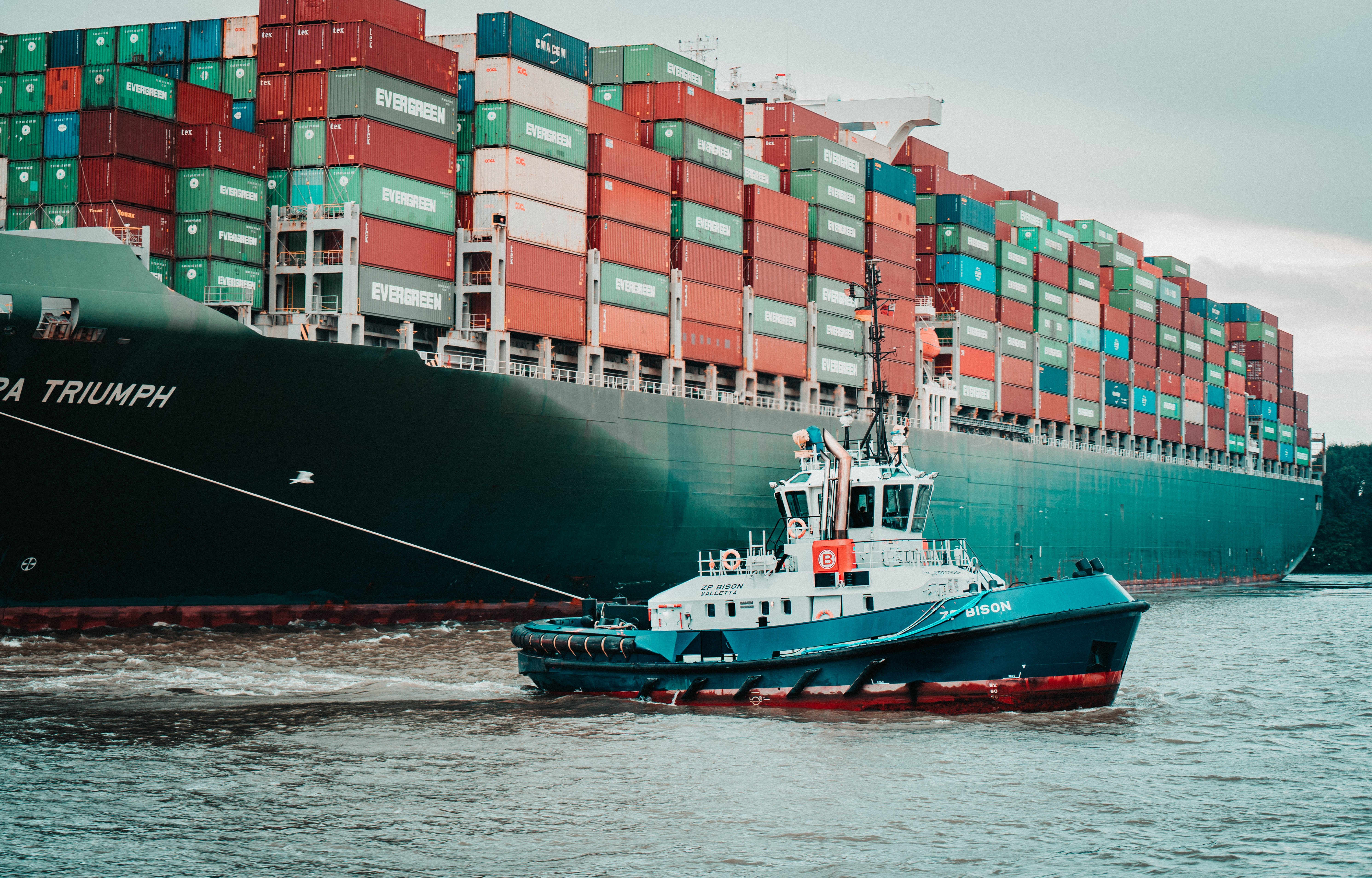 Das Bild zeigt die Fotografie eines sehr großen Frachtschiffes mit vielen Frachtcontainern, das von einem Schlepperboot in den Hafen gezogen wird.