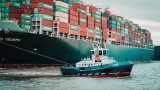 Das Bild zeigt die Fotografie eines sehr großen Frachtschiffes mit vielen Frachtcontainern, das von einem Schlepperboot in den Hafen gezogen wird.