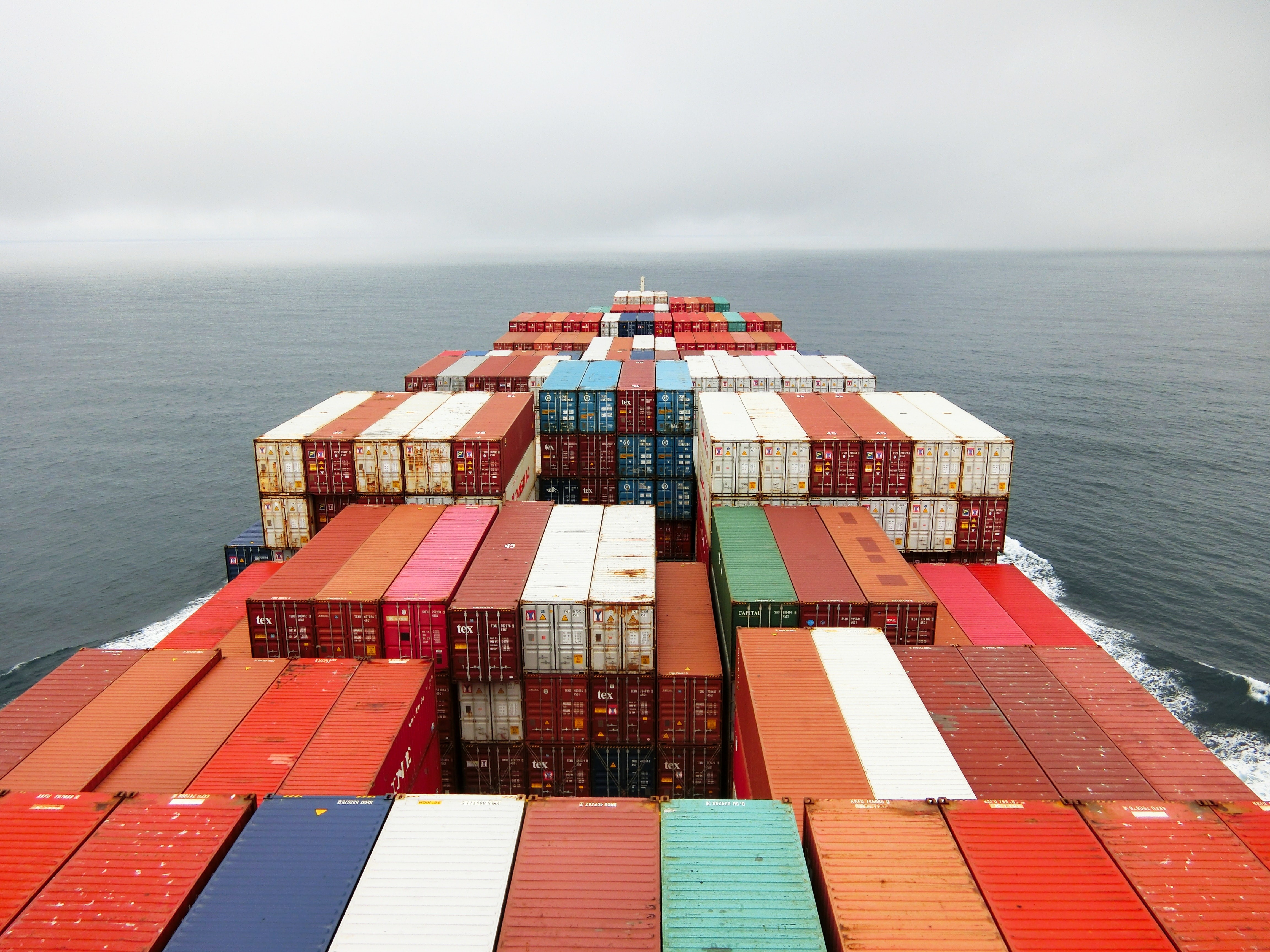 Das Bild zeigt die Fotografie eines Fachtschiffes von oben. Man sieht zahlreiche Frachtcontainer von oben. die auf einem Schiff verstaut sind, dass durch ein grau-blaues Meer auf den Horizont zufährt